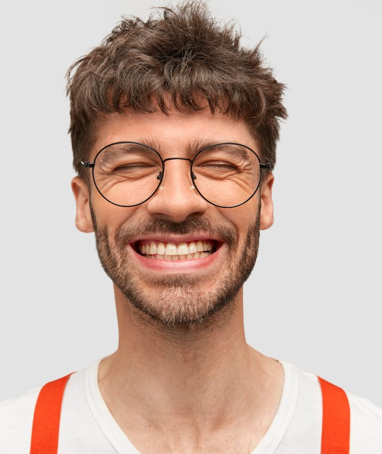 Positive bearded male hipster smiles broadly, has pleased expression, laughs at something funny, closes eyes, has trendy hairdo, isolated on white wall. Cheerful successful young businessman